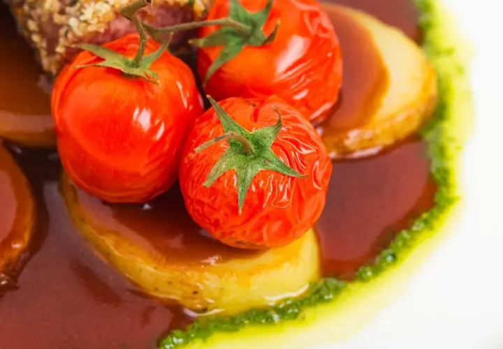 Pomodori alla griglia succosi su patate, immersi in una salsa scura e guarniti con un filo di pesto verde.