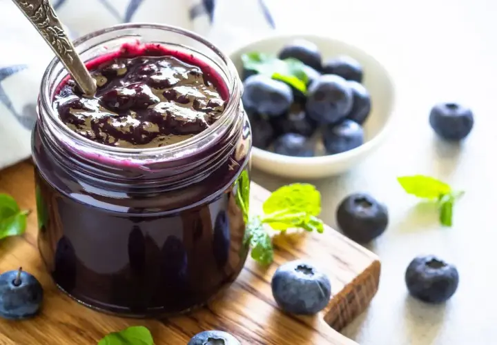 Vasetto di marmellata di mirtilli fatto in casa, con mirtilli freschi e foglie di menta su un tagliere di legno.