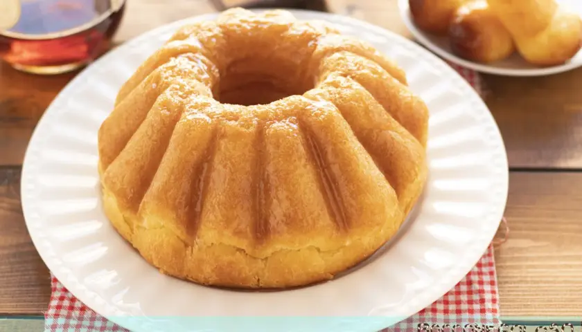 Un soffice babà a ciambella, lucido di bagna, pronto per essere gustato. Accanto, altri babà e una bottiglia di rum.