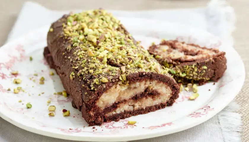 Rotolo al cioccolato e pistacchio, un dolce con farina di riso, ripieno di crema chiara e decorato con granella verde.