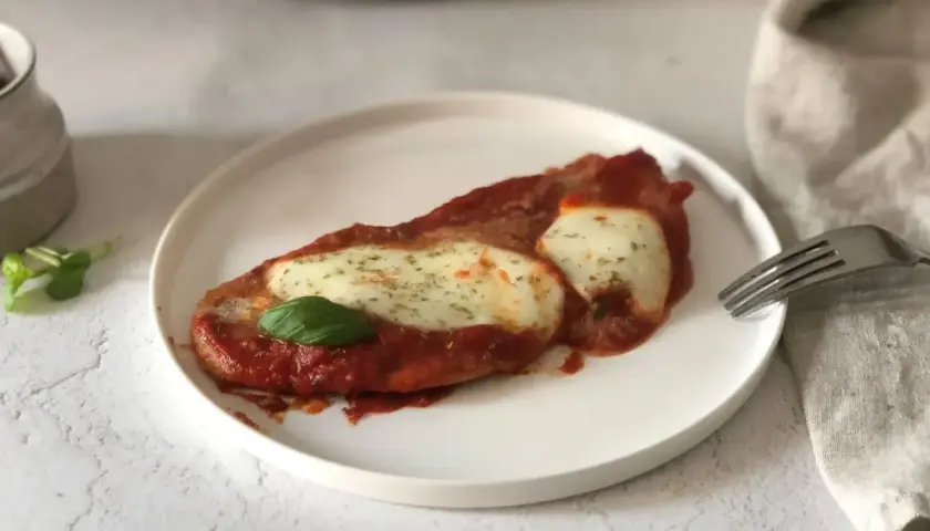 Scaloppine alla pizzaiola su un piatto bianco, con mozzarella filante, salsa di pomodoro e una foglia di basilico.