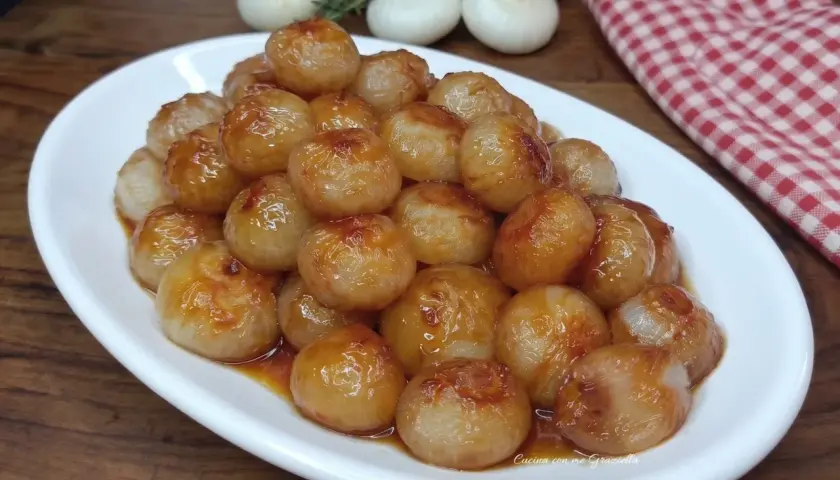 Cipolline in agrodolce, caramellate e lucide, servite su un piatto bianco. Sullo sfondo, cipolle fresche e un tovagliolo a quadri rossi.