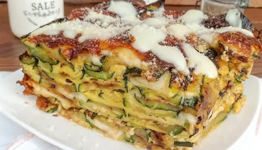 Una fetta di timballo di zucchine stratificato, con formaggio fuso e parmigiano grattugiato, servito su un piatto bianco.