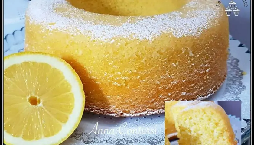 Un soffice ciambellone al limone, spolverato di zucchero a velo, accanto a una fetta di limone fresca.