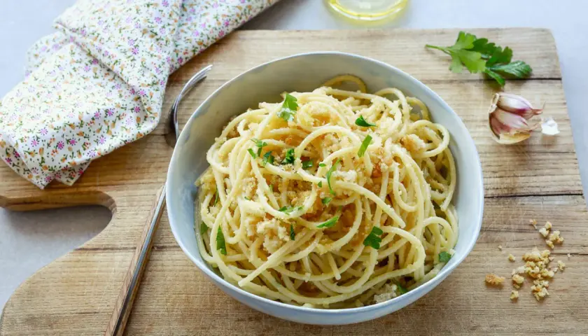 Un piatto di spaghetti con la mollica croccante, guarnito con prezzemolo fresco. Accanto, aglio e un bicchiere d'acqua.