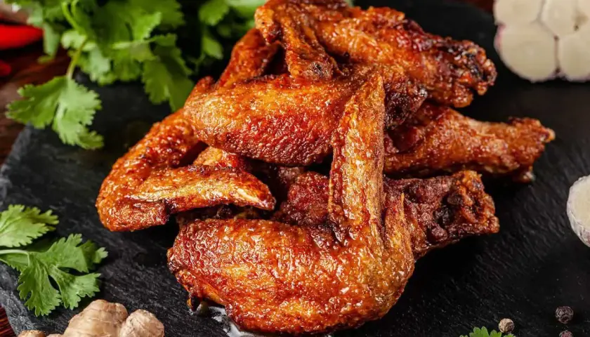 Un mucchio di ali di pollo dorate e succulente, condite con spezie, su una pietra scura, con coriandolo, zenzero e aglio sullo sfondo.