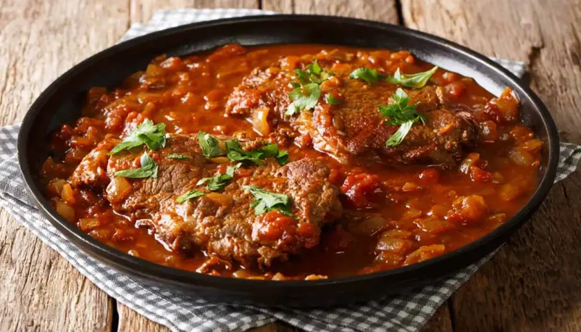 Carne alla pizzaiola morbida in un piatto scuro, con pomodoro e prezzemolo fresco.