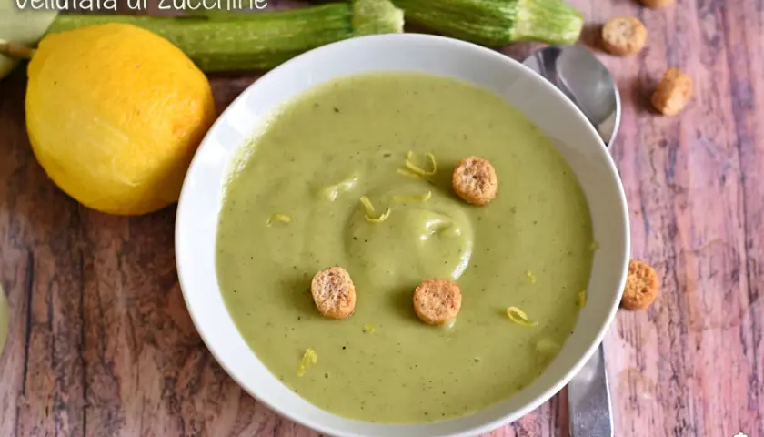 Vellutata di zucchine cremosa, guarnita con crostini e scorza di limone. Accanto, zucchine fresche e un limone intero.