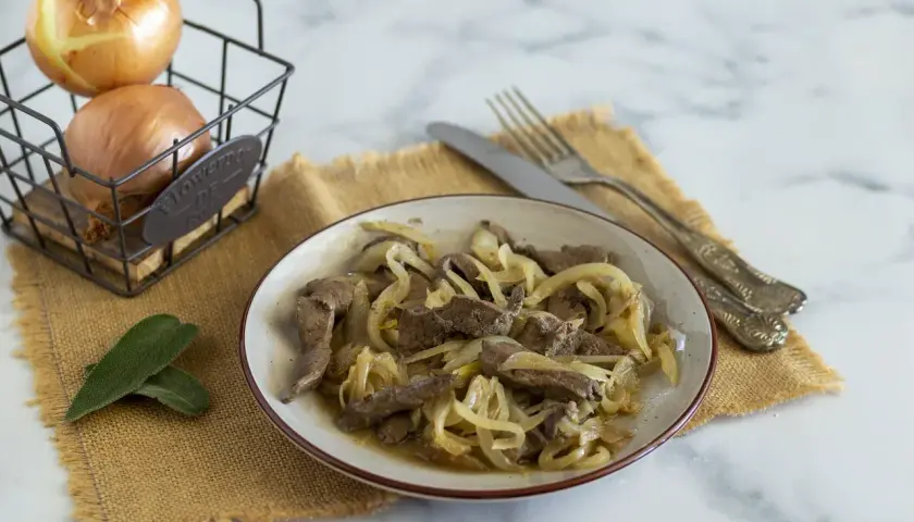 Piatto di fegato alla veneziana con cipolle dorate, accompagnato da due foglie di salvia e cipolle in un cestino.