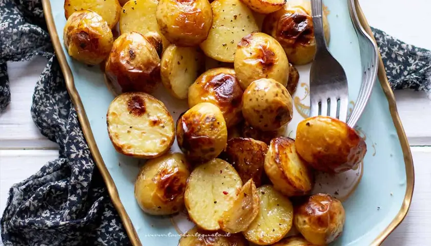 Patate novelle al forno dorate e croccanti, servite su un piatto azzurro con forchette.