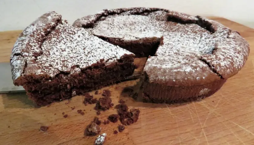 Una fetta di tenerina al cioccolato, cosparsa di zucchero a velo, è stata appena tagliata da una torta intera su un tagliere di legno.