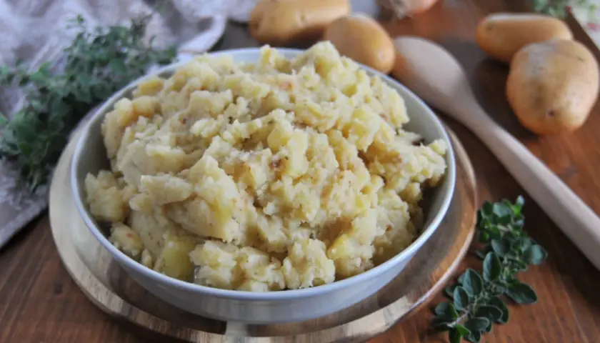 Patate in tecia, un contorno rustico e saporito, servito in una ciotola bianca su un tagliere di legno, con patate crude e erbe aromatiche sullo sfondo.