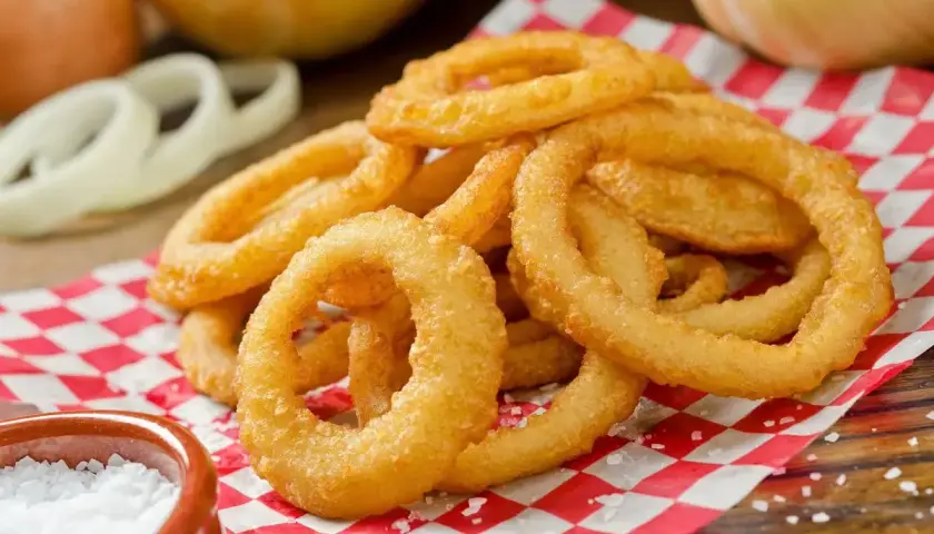 Anelli di cipolla dorati e croccanti, preparati con una **pastella per fritti** perfetta, serviti su carta a quadri rossi con sale grosso.