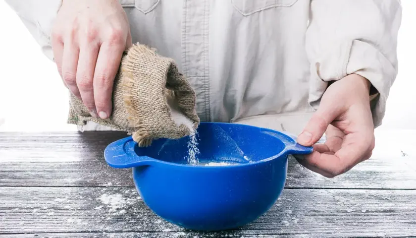 Mani versano zucchero da un sacchetto di juta in una ciotola blu, preparando ingredienti per pasta di zucchero.