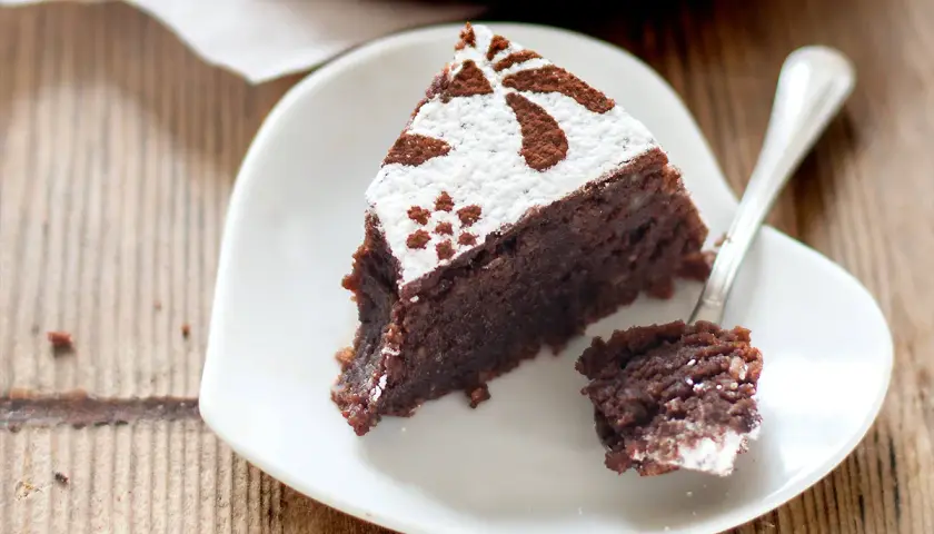 Una fetta di torta caprese bimby, soffice e al cioccolato, decorata con zucchero a velo e cacao, su un piatto bianco a forma di cuore.