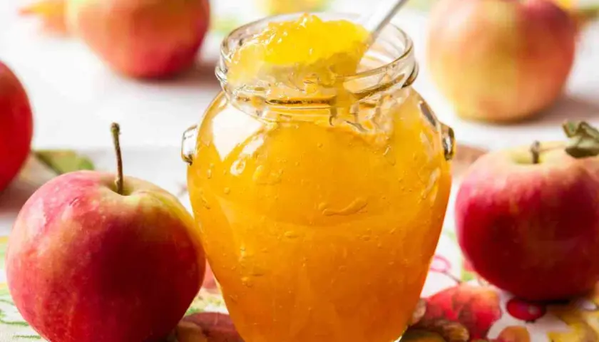 Un barattolo di marmellata di mele dorata, con una cucchiaiata che ne assaggia la dolcezza, circondato da mele rosse fresche.