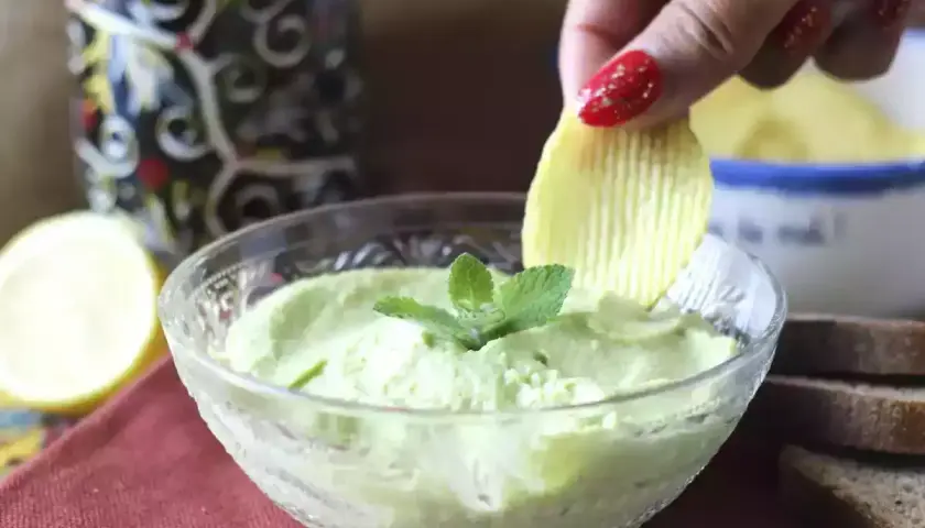 Una mano intinge una patatina in una cremosa maionese con uovo sodo, guarnita con menta.