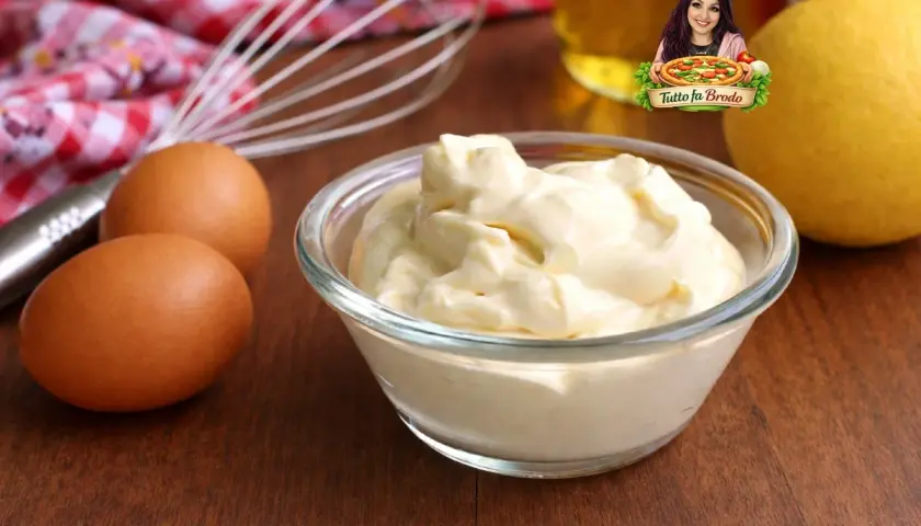 Una ciotola di cremosa maionese fatta in casa, con uova, limone e frusta sullo sfondo.