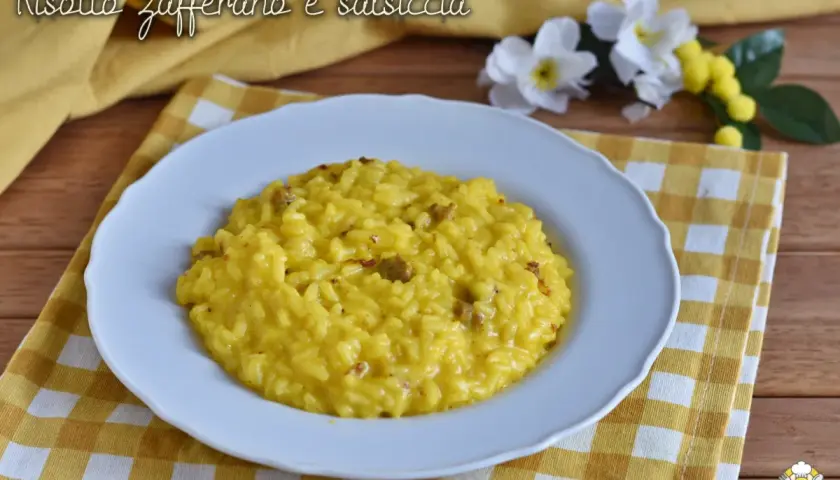 Un cremoso risotto salsiccia e zafferano, servito su un piatto bianco, con fiori gialli e bianchi sullo sfondo.