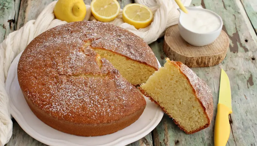 Deliziosa torta per colazione, soffice e profumata al limone, spolverata di zucchero a velo, con una fetta tagliata e una ciotolina di yogurt.