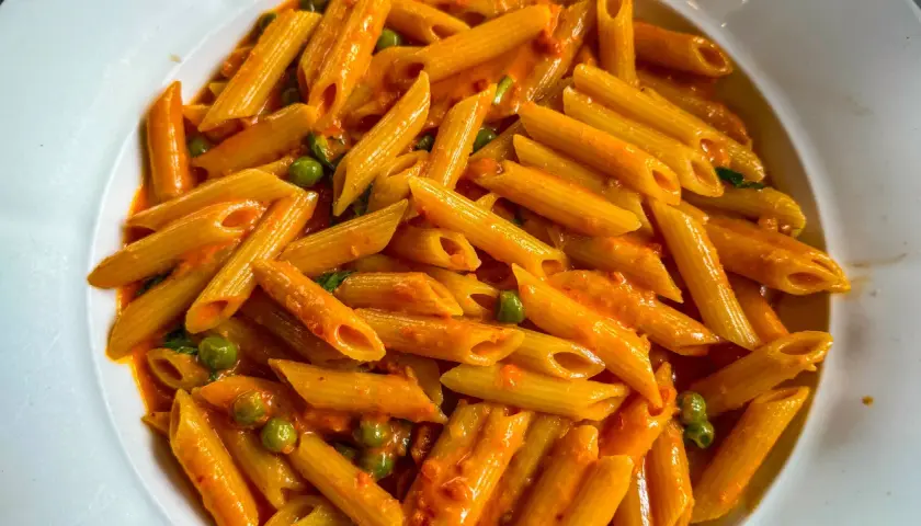 Penne ai 7 peccati, un piatto di pasta avvolto in una cremosa salsa rossa, con piselli verdi che spuntano come gemme preziose.