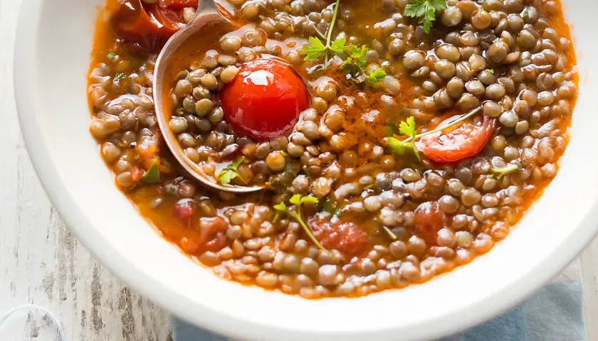 Una ciotola di zuppa di lenticchie fumante, con pomodorini e prezzemolo fresco. Un cucchiaio raccoglie una porzione generosa di questo piatto confortante.