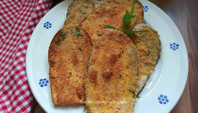 Fette dorate di cotoletta di melanzane impanate, guarnite con menta fresca, su un piatto bianco con decori blu.