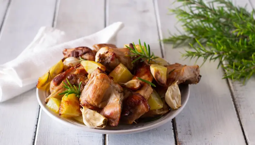 Piatto invitante di coniglio al forno con patate arrosto, aglio e rosmarino su un tavolo di legno.
