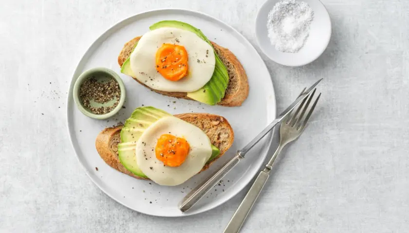 Due fette di pane tostato con avocado, formaggio e un occhio di bue, condite con pepe.