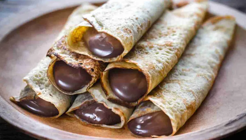 Un mucchio di crepes arrotolate, ripiene di golosa Nutella, su un piatto di legno.