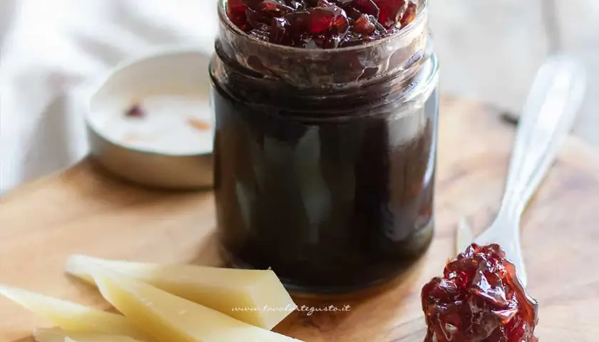 Un vasetto di marmellata di cipolle rosse, con un cucchiaino che ne raccoglie una porzione, accanto a fette di formaggio su un tagliere di legno.