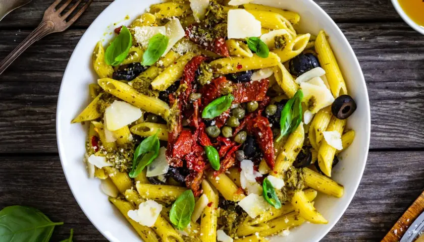 Penne con pesto, pomodori secchi, olive nere, capperi e scaglie di parmigiano. Un piatto ricco di sapori mediterranei.