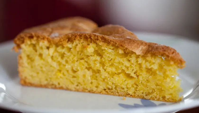 Una fetta di torta alle mandorle dorata e soffice, con una crosta leggermente caramellata, servita su un piatto bianco.