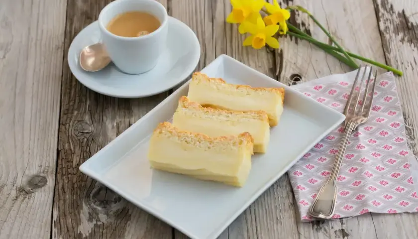 Tre fette di torta magica ricetta, cremosa e dorata, su un piatto bianco, con caffè e narcisi.