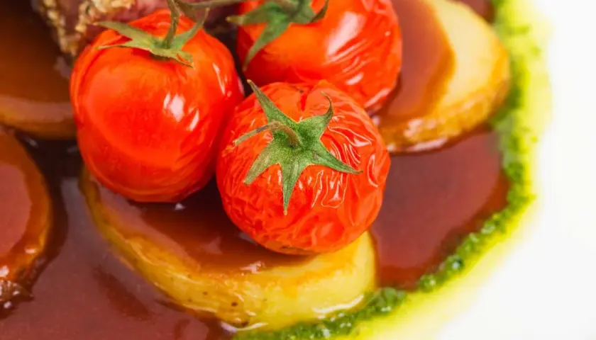 Pomodori alla griglia succosi su patate, immersi in una salsa scura e guarniti con un filo di pesto verde.