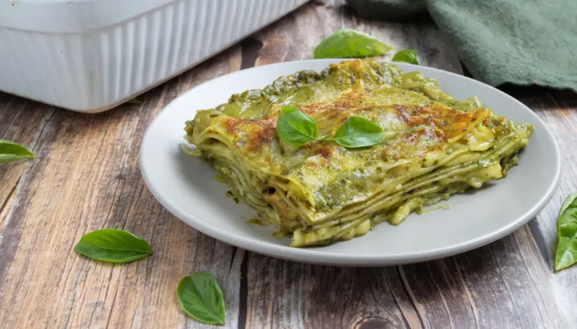 Una fetta di lasagne al pesto, stratificata e dorata, guarnita con basilico fresco su un piatto grigio.