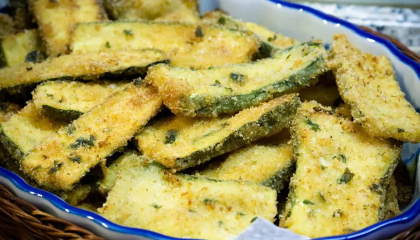 Fettine di zucchine impanate e dorate, appena tolte dalla padella, servite in una ciotola.