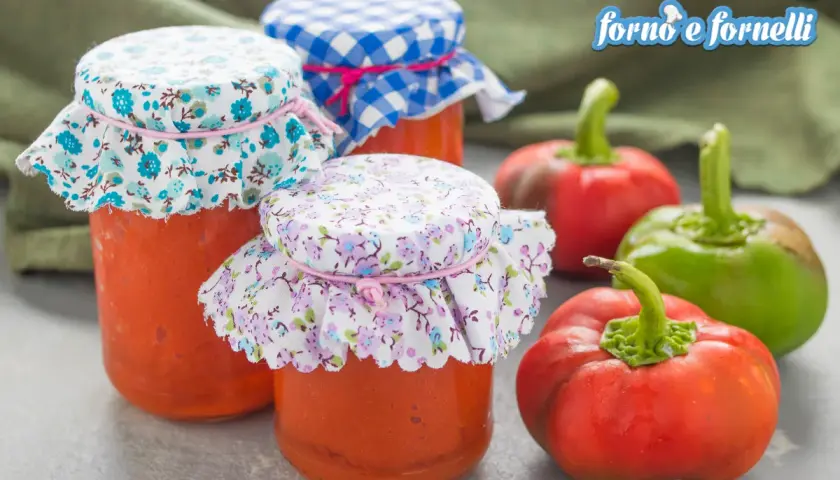 Barattoli di crema di peperoni fatti in casa, decorati con stoffe floreali e a quadretti, accanto a peperoni freschi.