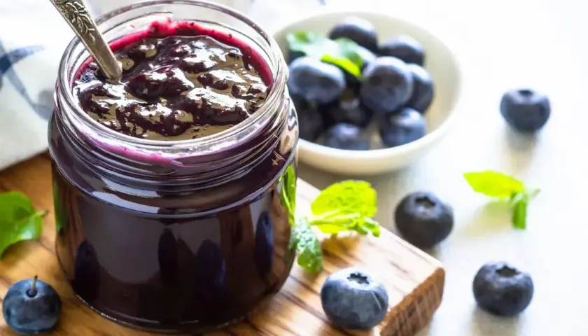 Vasetto di marmellata di mirtilli fatto in casa, con mirtilli freschi e foglie di menta su un tagliere di legno.