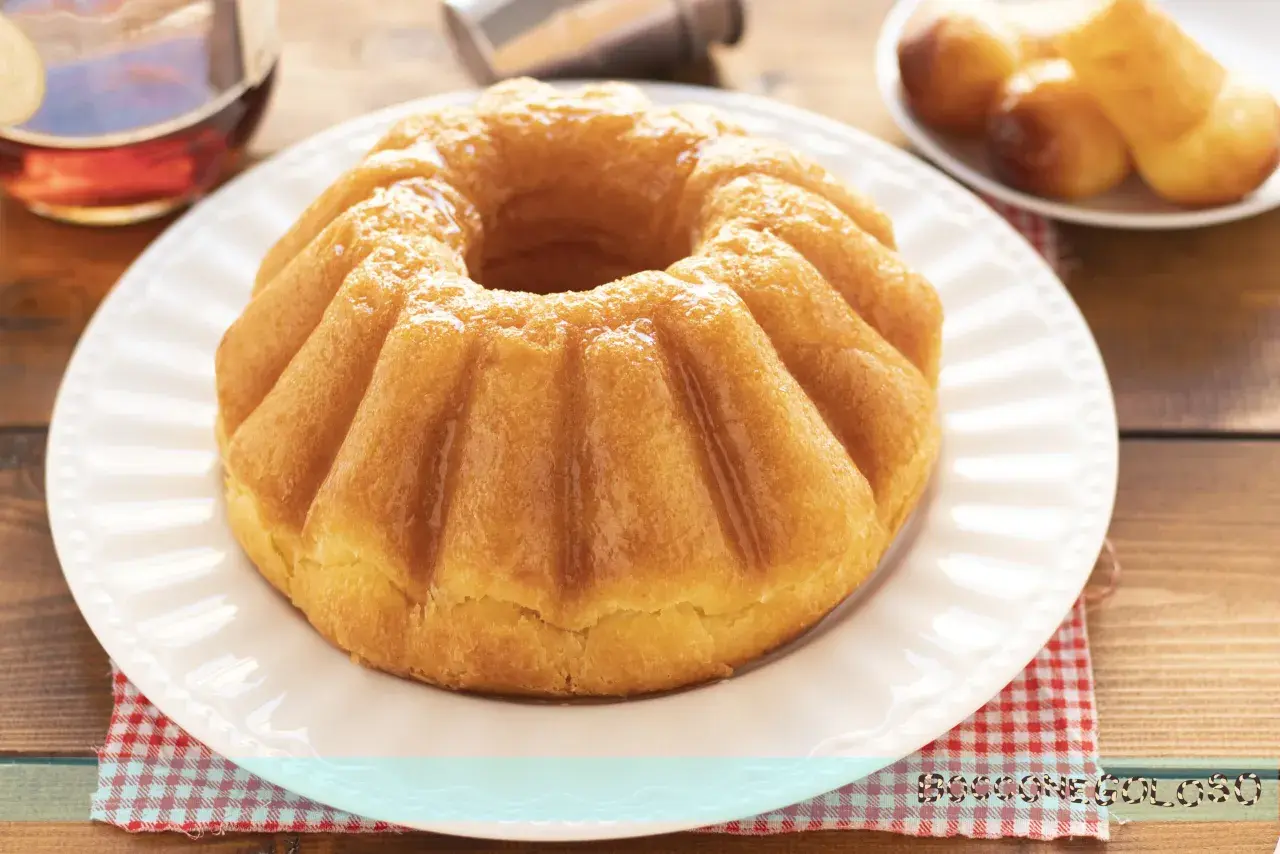 Un soffice babà a ciambella, lucido di bagna, pronto per essere gustato. Accanto, altri babà e una bottiglia di rum.