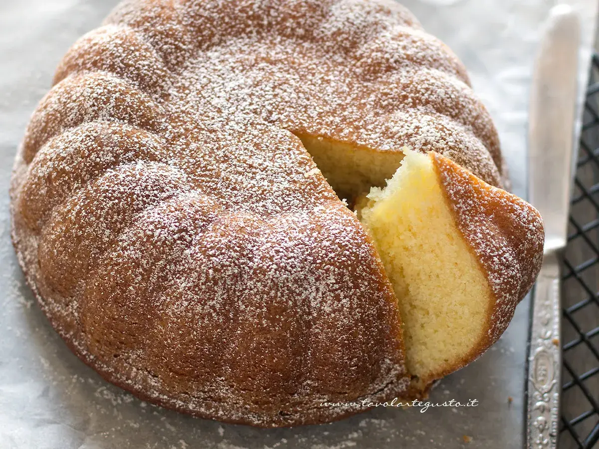 Una soffice torta allo yogurt, cosparsa di zucchero a velo, con una fetta tagliata che ne rivela l'interno dorato e invitante.