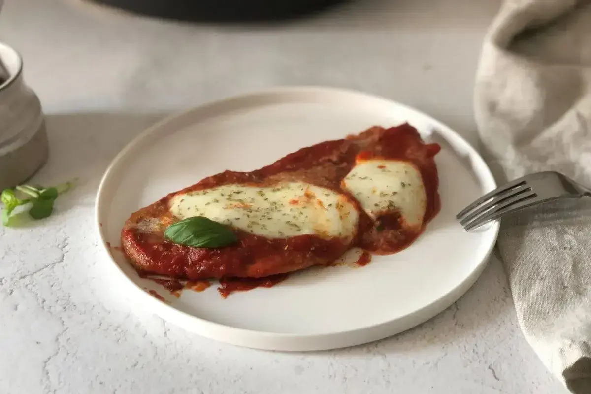 Scaloppine alla pizzaiola su un piatto bianco, con mozzarella filante, salsa di pomodoro e una foglia di basilico.