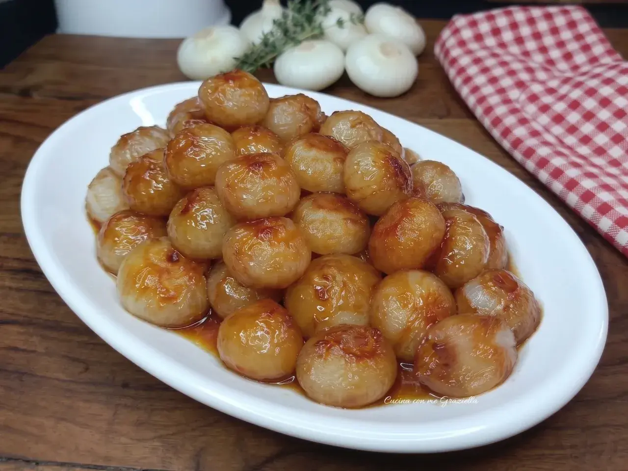 Cipolline in agrodolce, caramellate e lucide, servite su un piatto bianco. Sullo sfondo, cipolle fresche e un tovagliolo a quadri rossi.