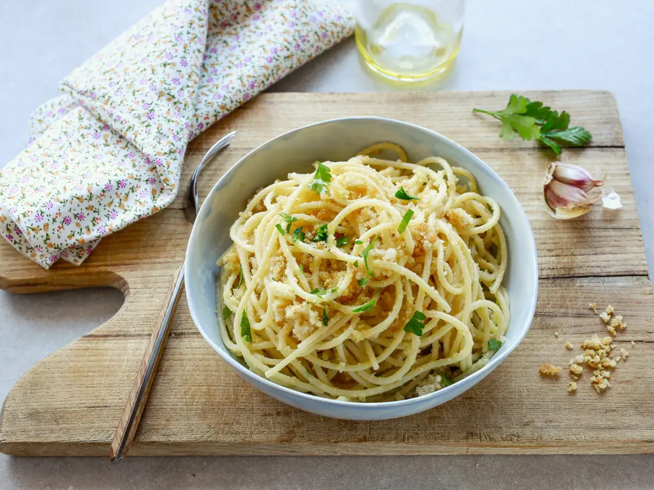 Un piatto di spaghetti con la mollica croccante, guarnito con prezzemolo fresco. Accanto, aglio e un bicchiere d'acqua.
