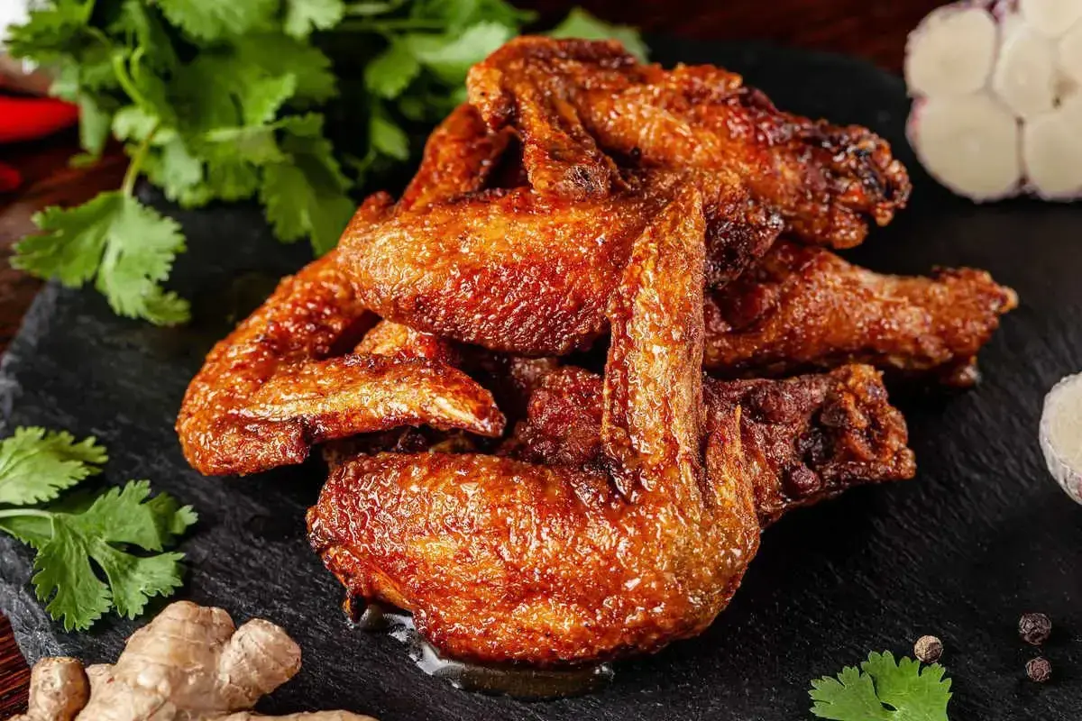 Un mucchio di ali di pollo dorate e succulente, condite con spezie, su una pietra scura, con coriandolo, zenzero e aglio sullo sfondo.