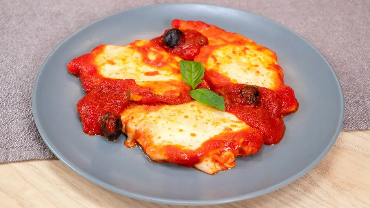 Delizioso pollo alla pizzaiola su un piatto grigio, con pomodoro, mozzarella filante, olive nere e una foglia di basilico.