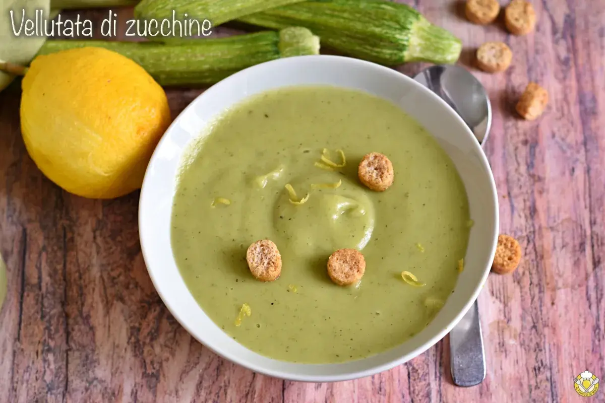 Vellutata di zucchine cremosa, guarnita con crostini e scorza di limone. Accanto, zucchine fresche e un limone intero.