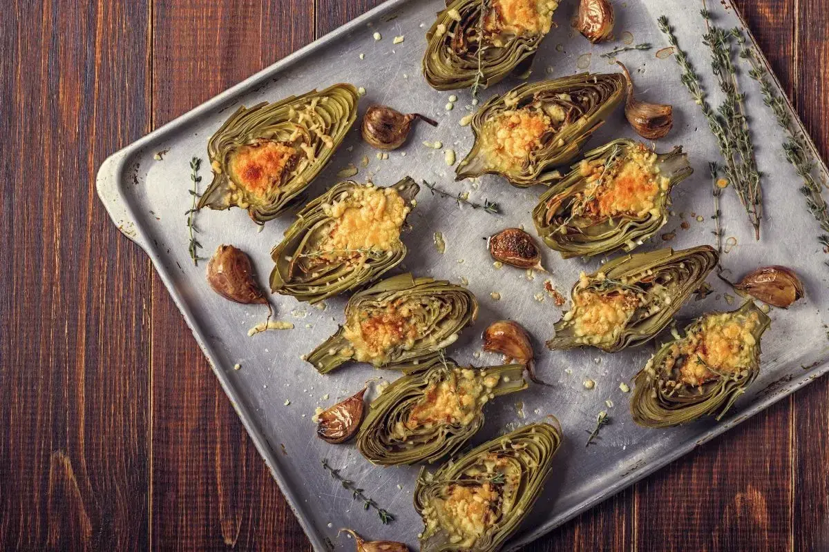 Carciofi al forno morbidi, ripieni e gratinati, con aglio e timo su teglia metallica.