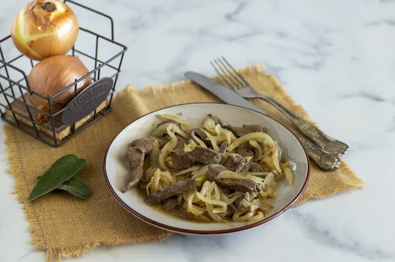 Piatto di fegato alla veneziana con cipolle dorate, accompagnato da due foglie di salvia e cipolle in un cestino.
