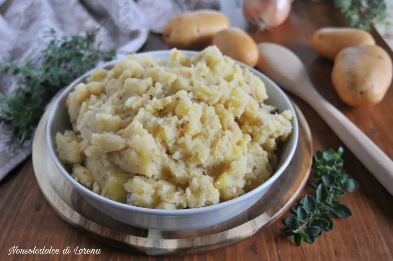 Patate in tecia, un contorno rustico e saporito, servito in una ciotola bianca su un tagliere di legno, con patate crude e erbe aromatiche sullo sfondo.